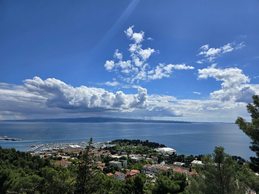 Pogled s Marjana pruža se na splitski poluotok, gradsku lučku zonu i Kaštelanski zaljev s otokom Čiovom i Bračem u pozadini pod vedrim plavim nebom.